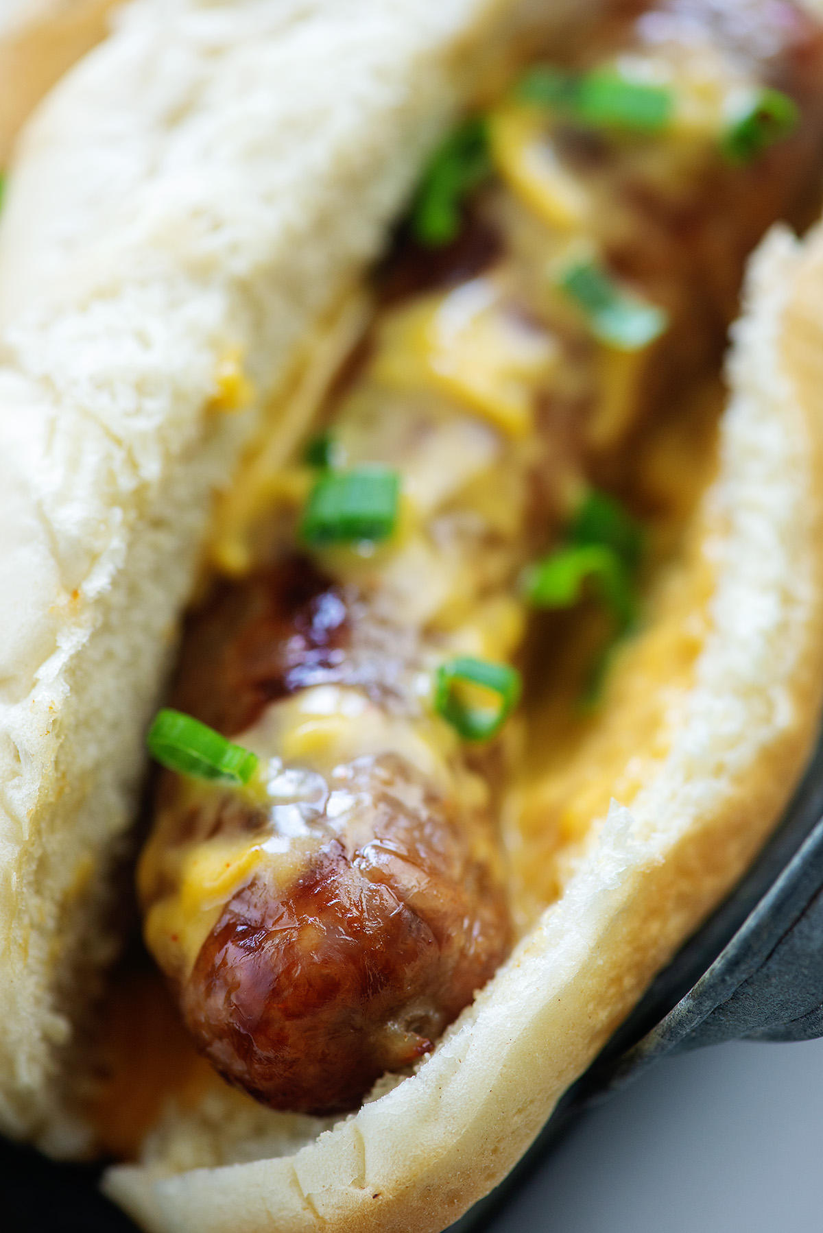 A close up of a bratwurst on a bun topped with beer cheese sauce and diced green onions.