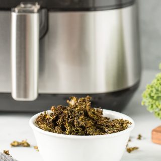 Kale chips in a white bowl in front of an air fryer.