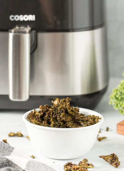 Kale chips in a white bowl in front of an air fryer.