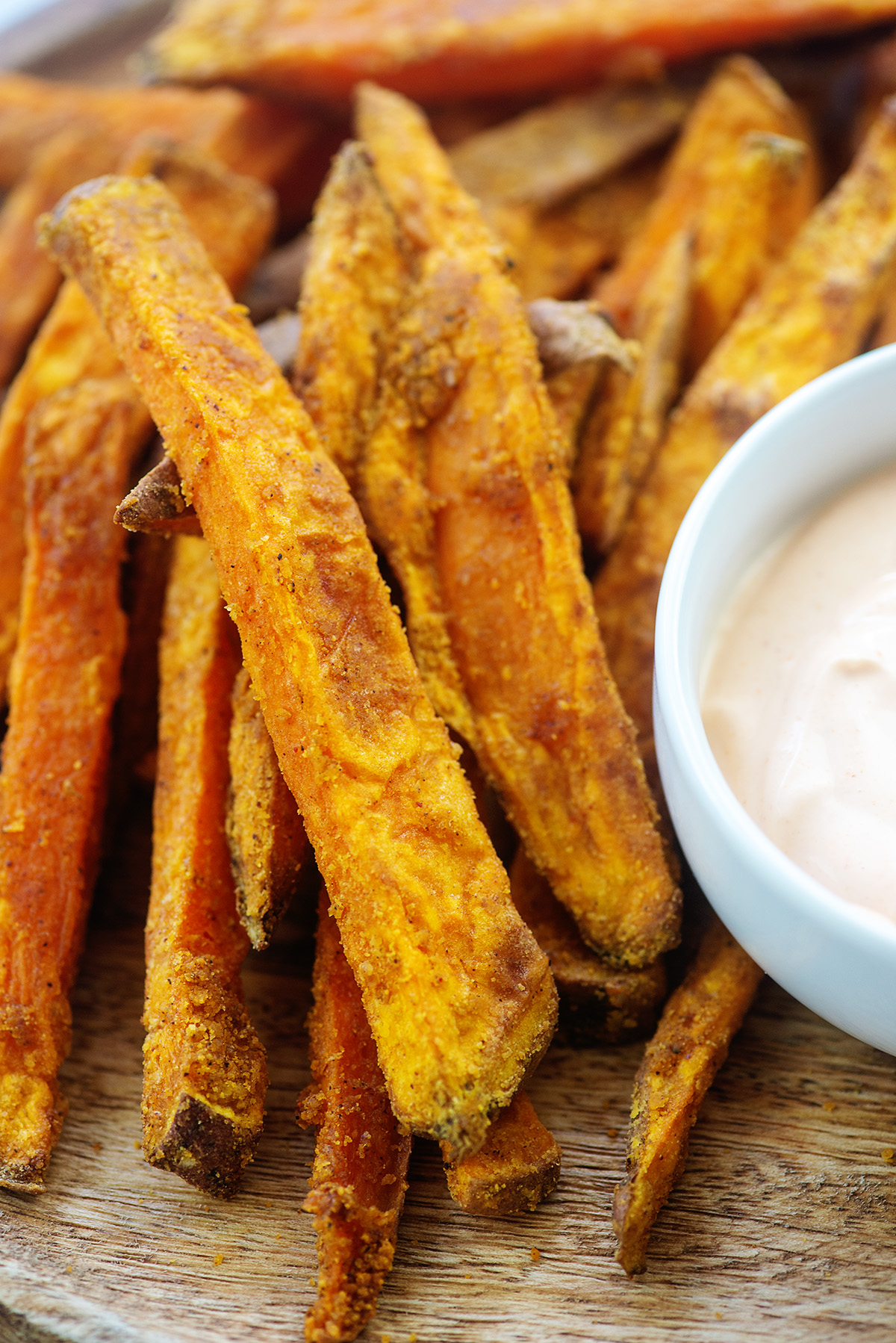 A close up of sweet potato fries on a wooden cutting board next to a bowl of dip.