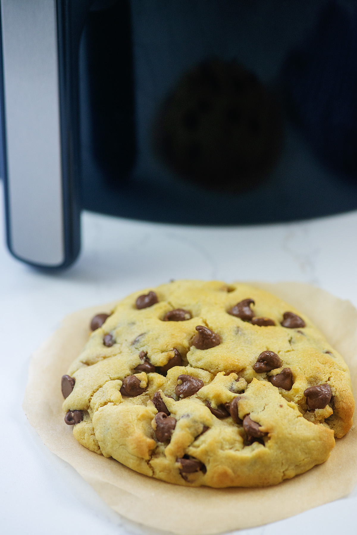 A single chocolate chip cookie in front of an air fryer