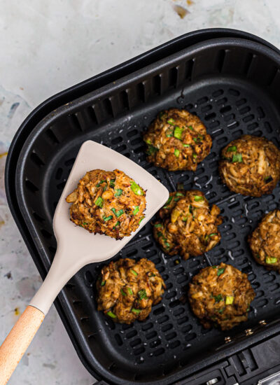 a spatula holding a cooked crab cake above an air fryer basket of crab cakes.