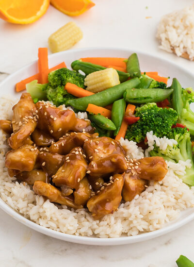 A plate of orange chicken over rice next to a pile of vegetables.