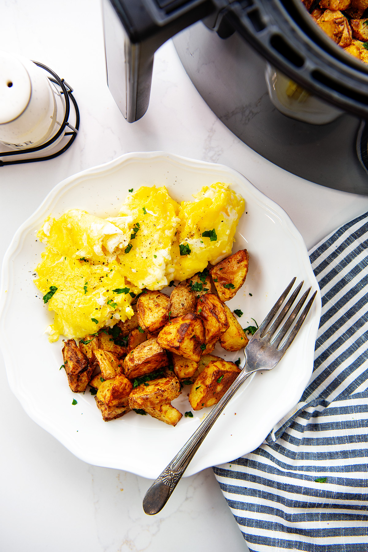 Overhead view of eggs and potatoes on a small white plate next to an air fryer basket.