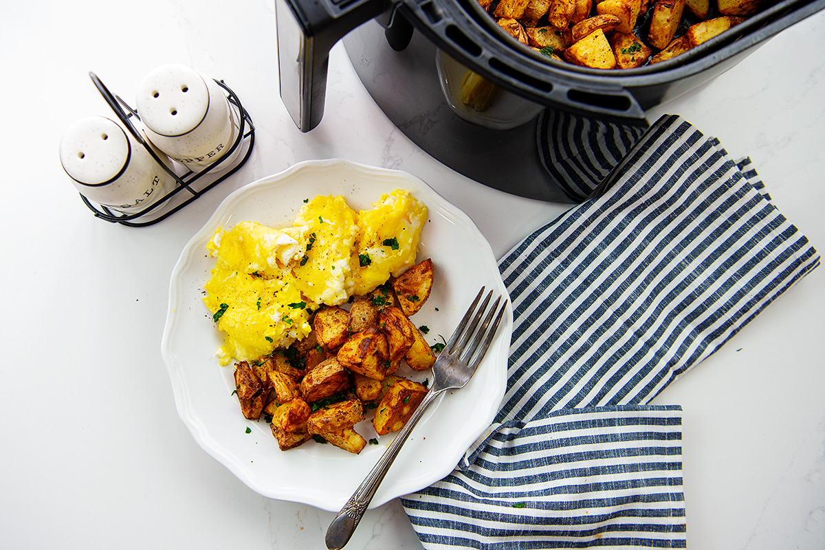 Overhead view of breakfast potatoes and eggs on a white plate next to an air fryer basket.