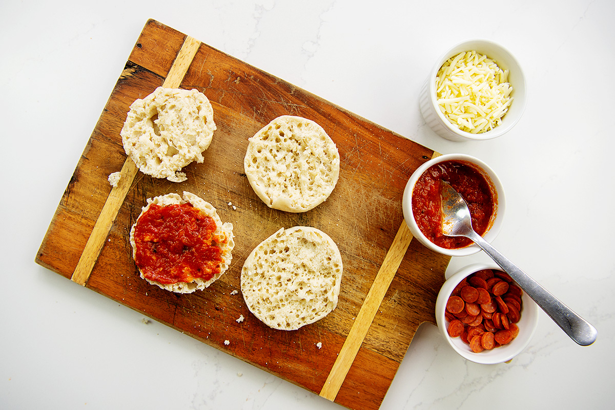 Overhead view of an English1 muffin on a cutting board next to cups of pizza toppings.