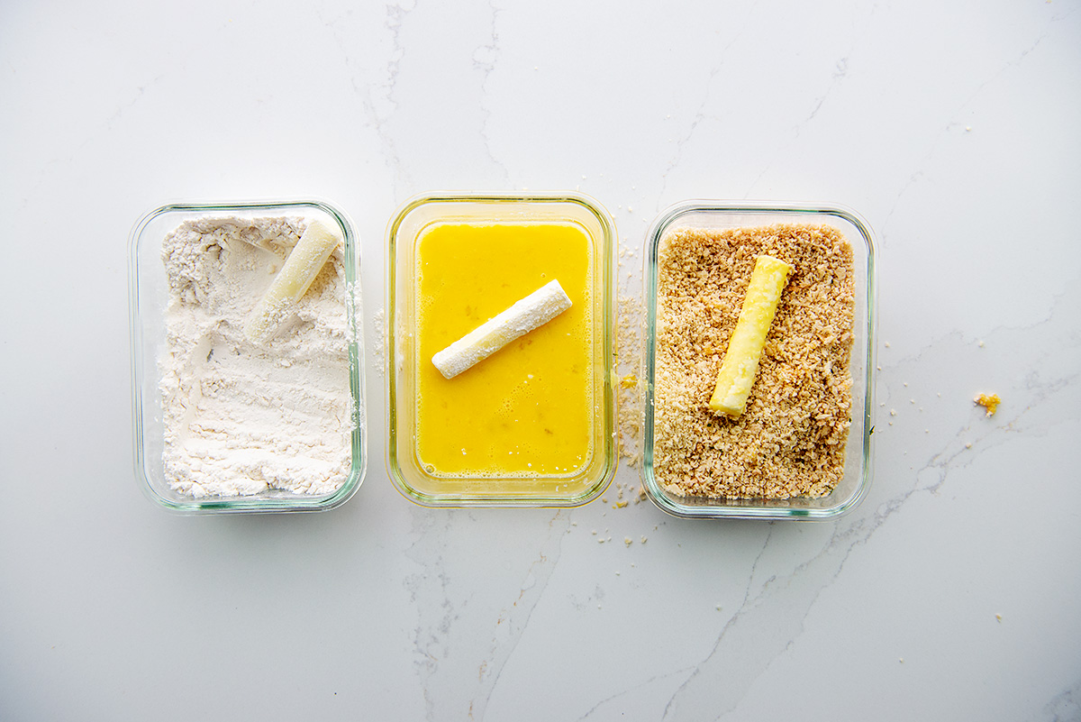 Three shallow dishes of cheese sticks in different stages of breading.