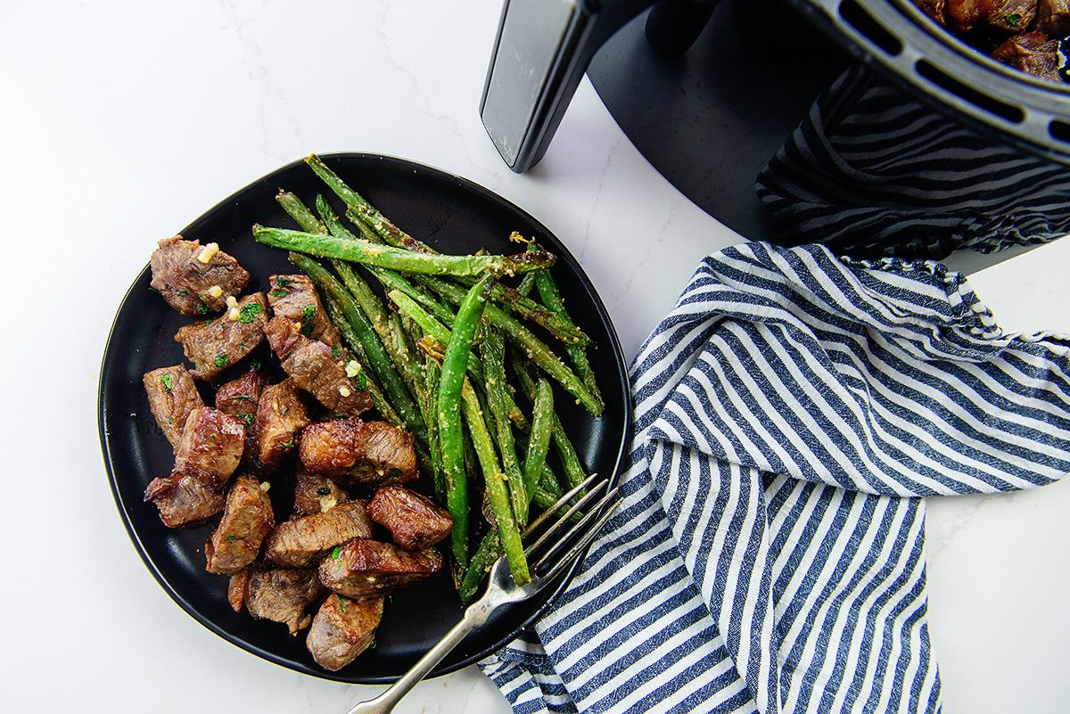Overhead view of steak bites and asparagus on a plate in front of an air fryer basket
