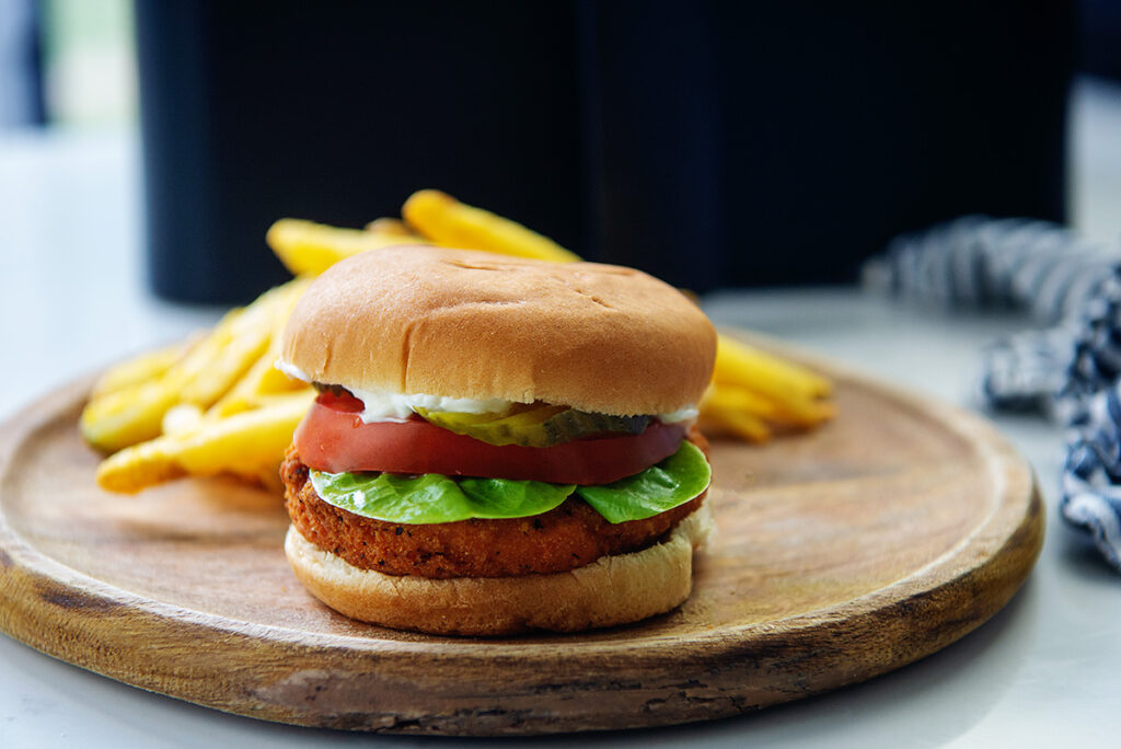 Chicken sandwich on a wooden plate in front of a pile of fries