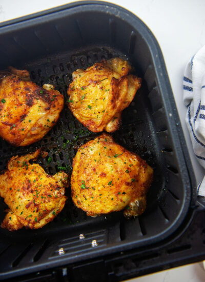 Overhead view of four cooked chicken thighs in an air fryer basket on a white counter top.