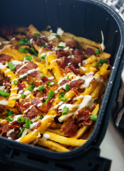 Loaded fries in an air fryer basket with bacon, cheese, ranch, and green onions.