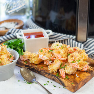 Coconut shrimp piled up on a cutting board in front of an air fryer.