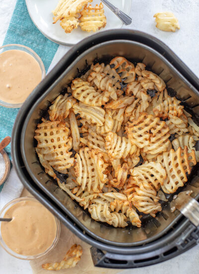Overhead view of waffle fries in an air fryer surrounded by fry sauce cups.