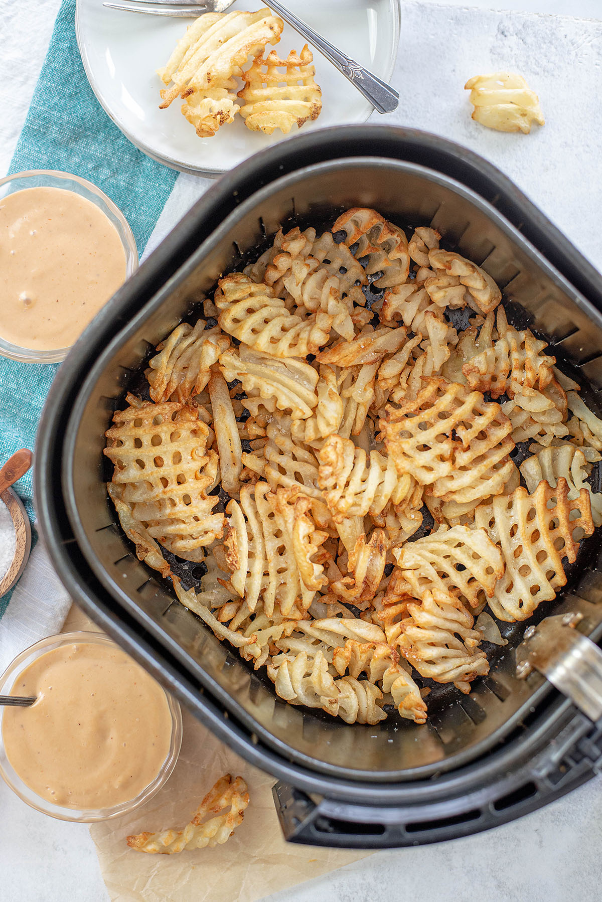 Overhead view of waffle fries in an air fryer surrounded by fry sauce cups.