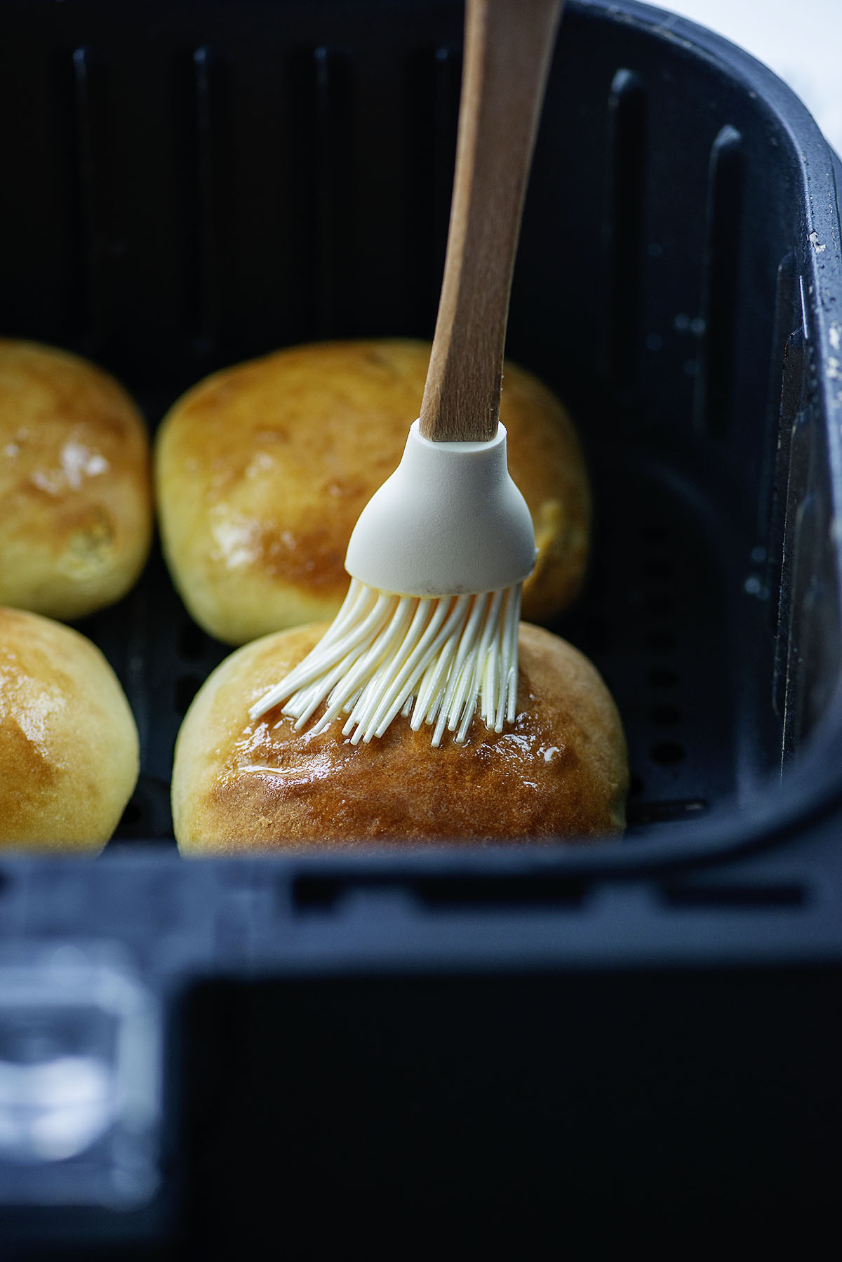 Brushing butter on top of a dinner roll in an air fryer basket.