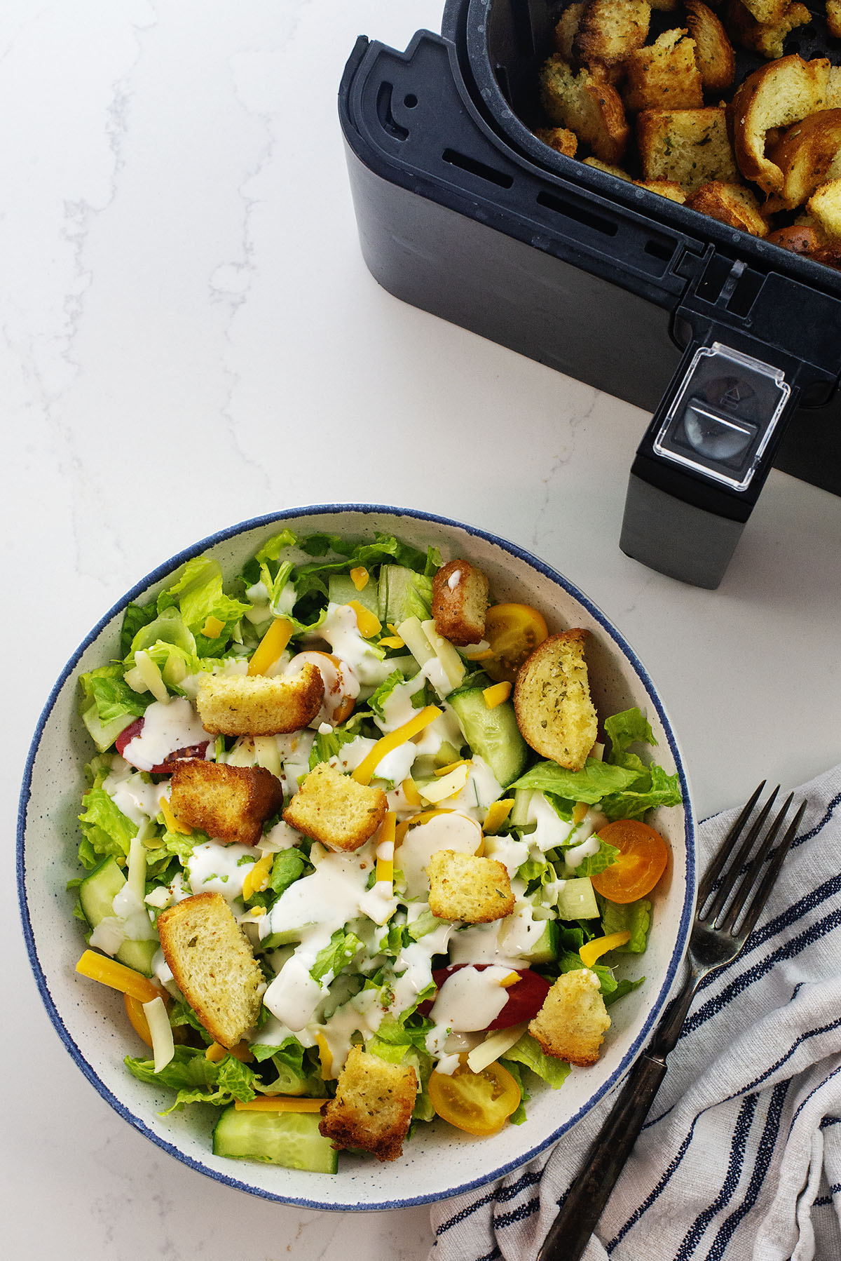 Overhead view of a salad with croutons in front of an air fryer.