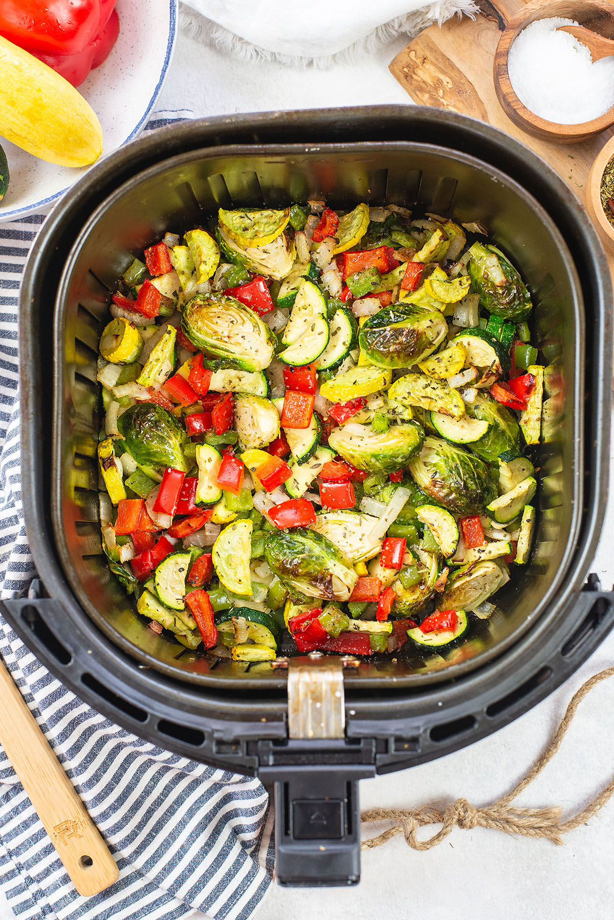 Overhead view of an air fryer basket full of air fried vegetables surrounded by vegetables.