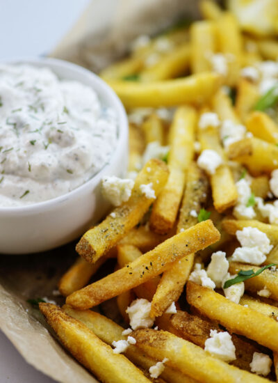 French fries with feta cheese and seasoning on them with a cup of tatziki sauce.