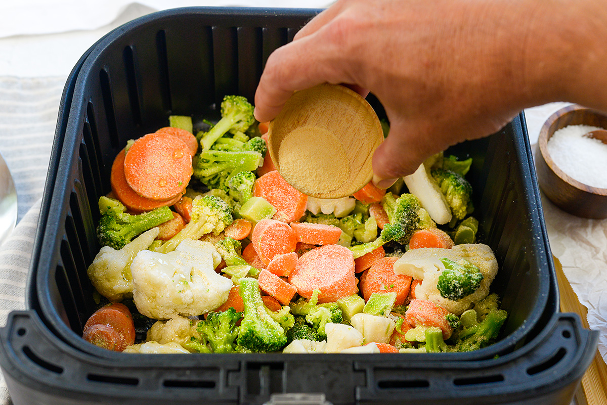 Person adding seasoning to veggies in an air fryer basket.