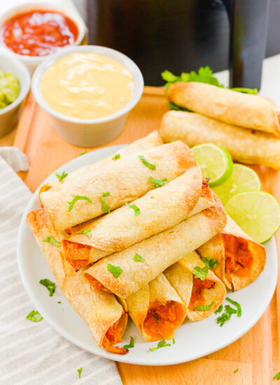 Chicken taquitos stacked on a plate in front of an air fryer basket.