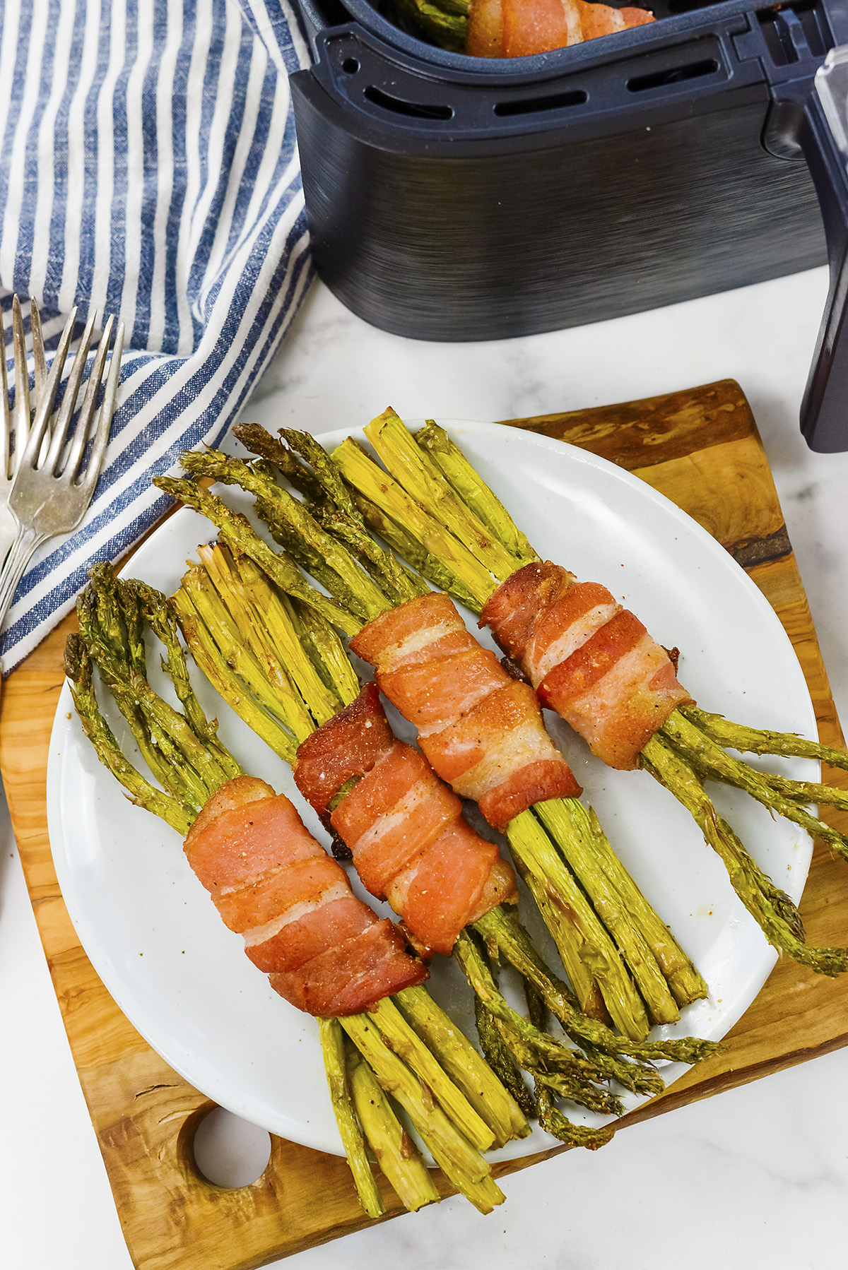 Bacon wrapped asparagus on a white plate in front of an air fryer.