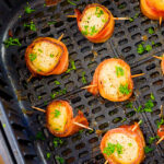 Several bacon wrapped scallops in an air fryer basket.