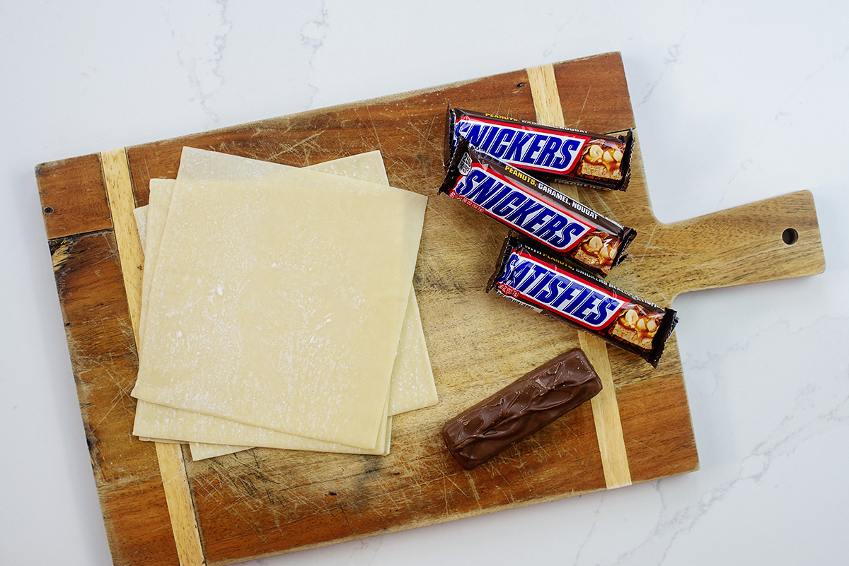 Egg roll wrappers and snickers bars on a wooden cutting board.