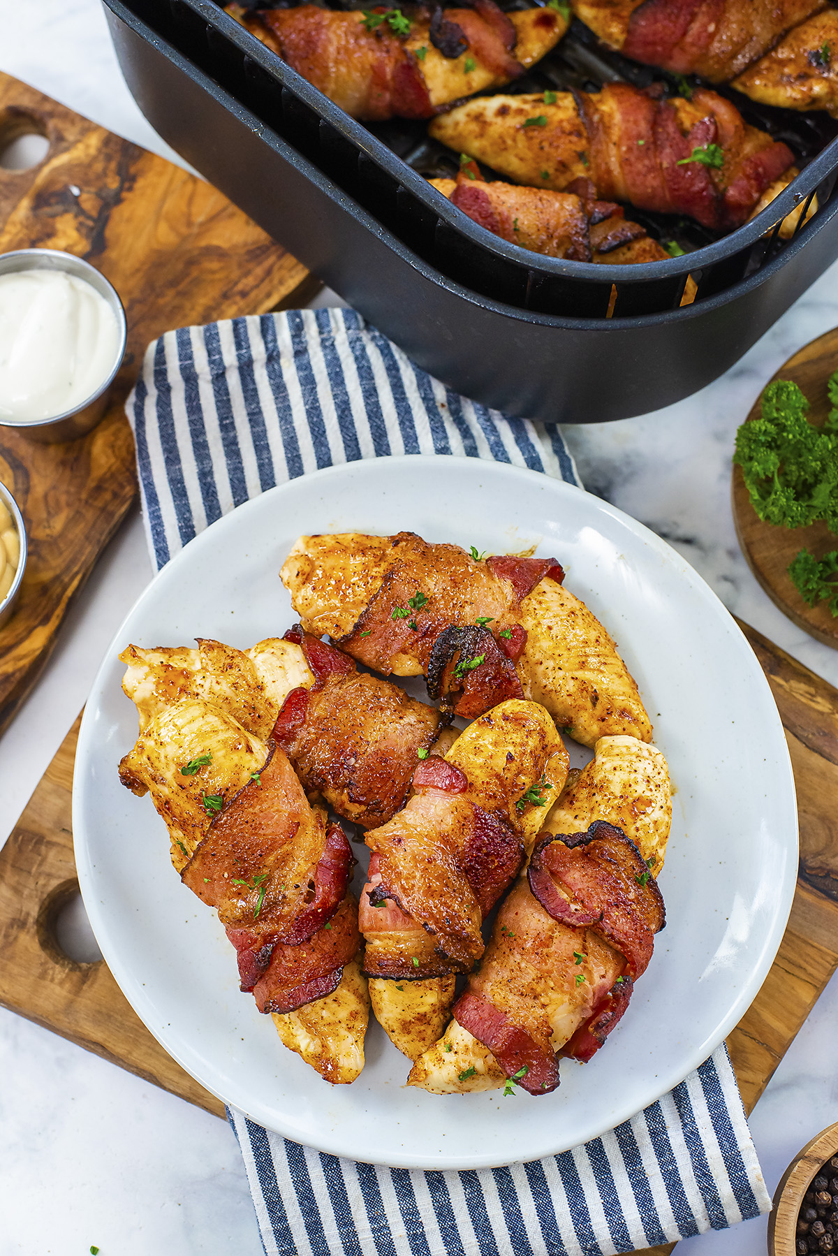 Bacon wrapped chicken tenders on a plate in front of an air fryer basket.