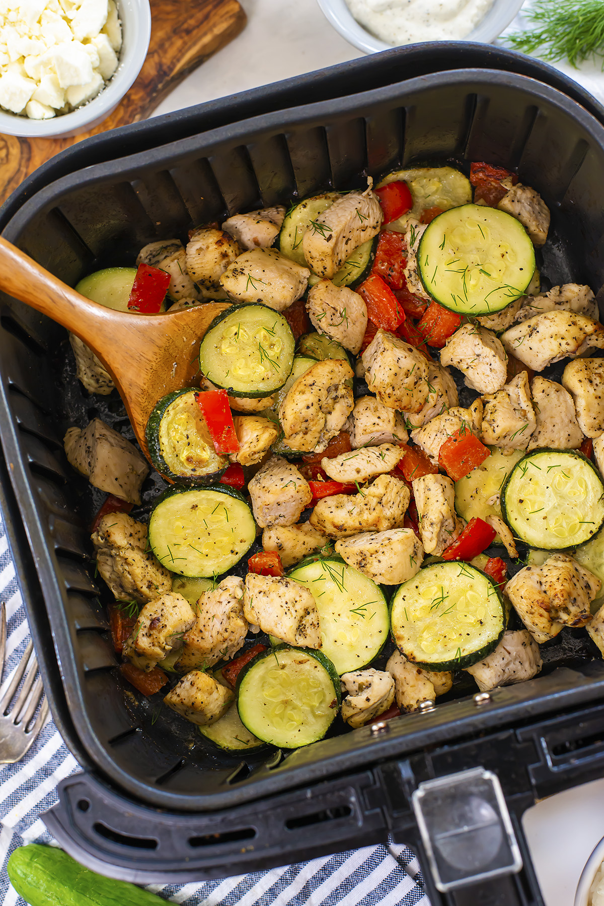 A wooden spoon in an air fryer basket full of Greek chicken, zucchini, and red peppers.