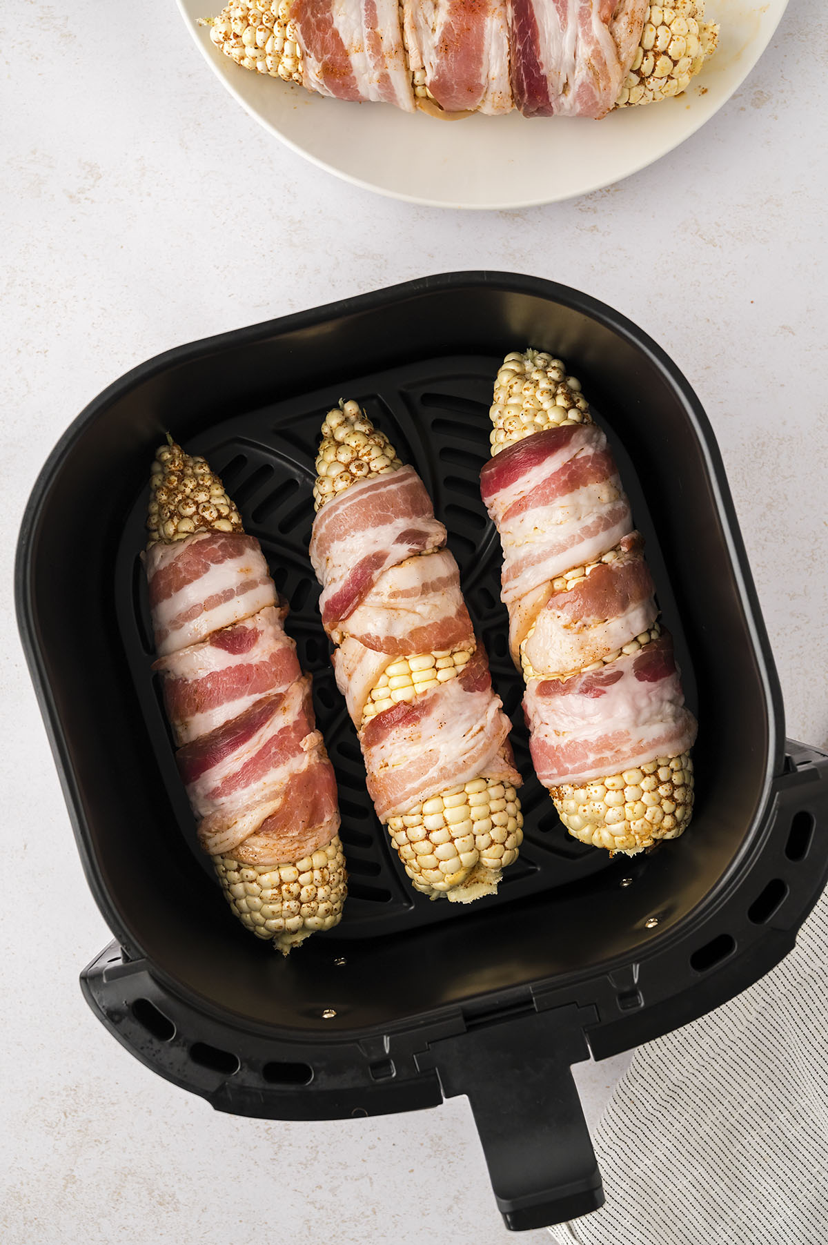 Three raw bacon wrapped corn on the cobs in an air fryer basket.