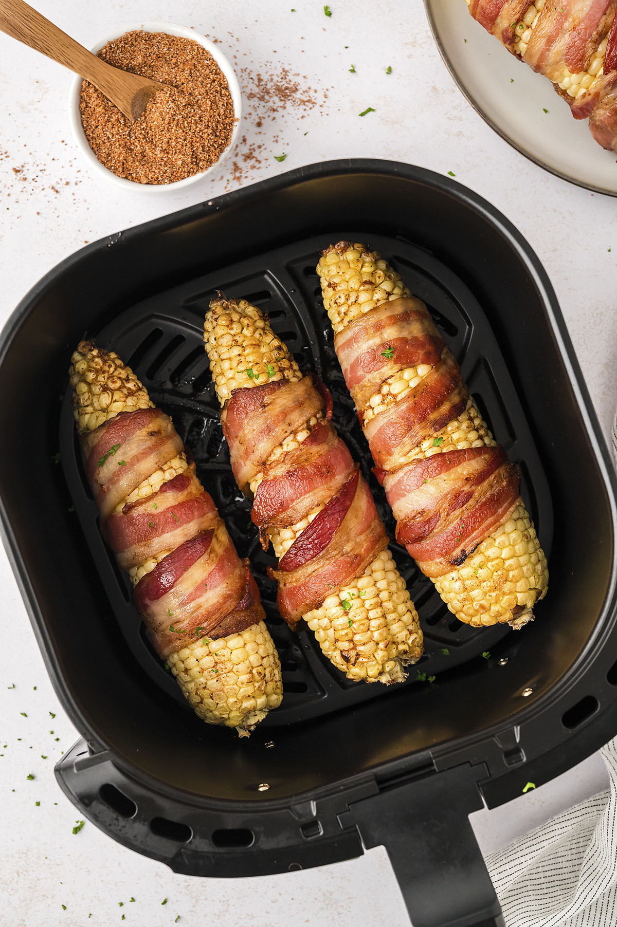 Three bacon wrapped corn on the cobs in an air fryer basket.