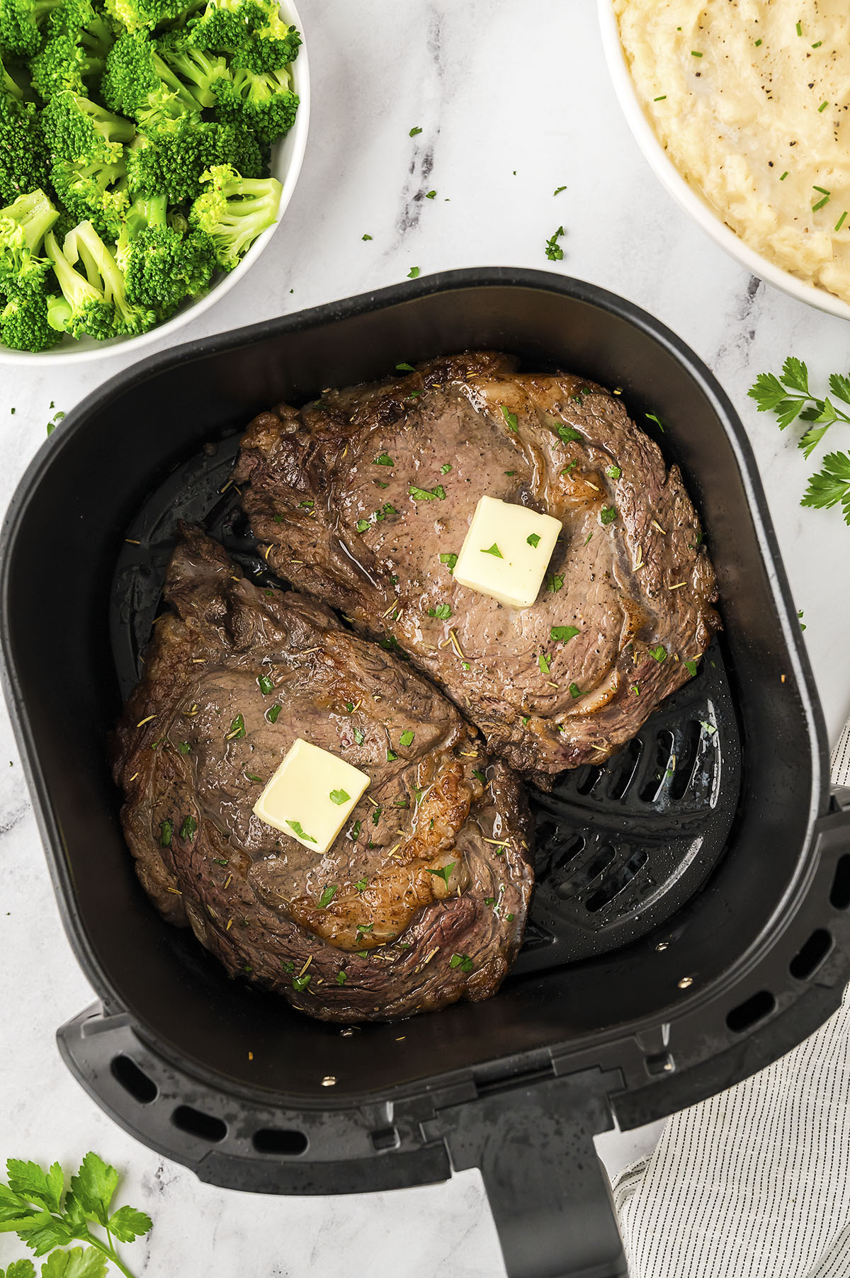Two ribeyes with butter in an air fryer basket.