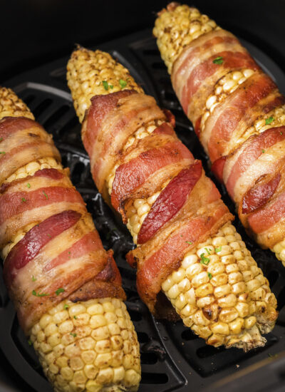 Close up of three bacon wrapped corn cobs in an air fryer basket.