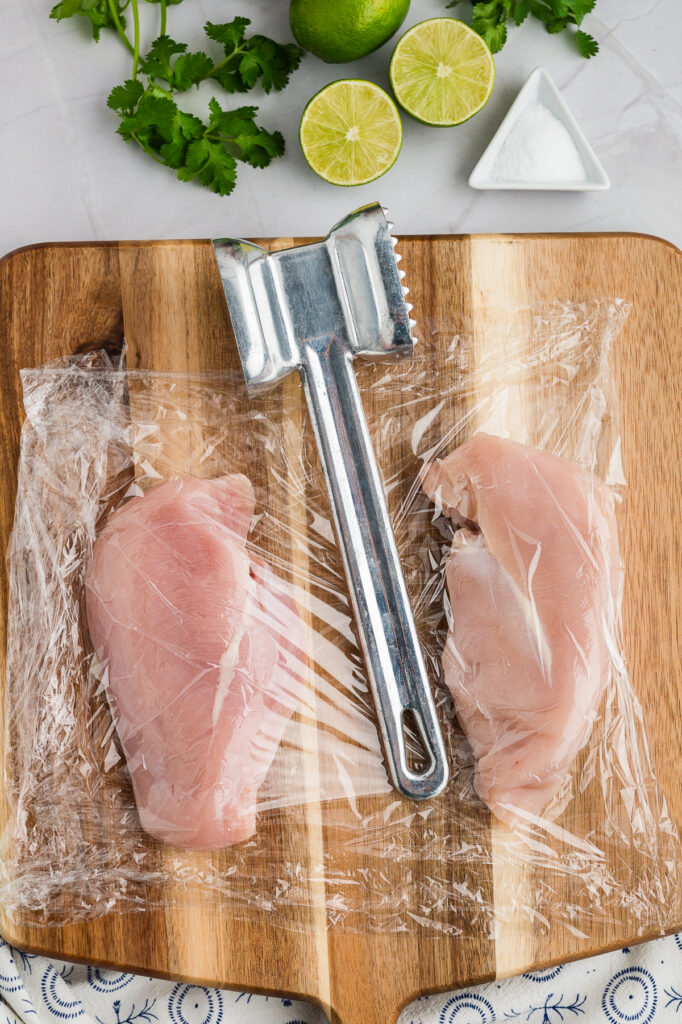 A tenderizer and raw chicken breast on a cutting board.