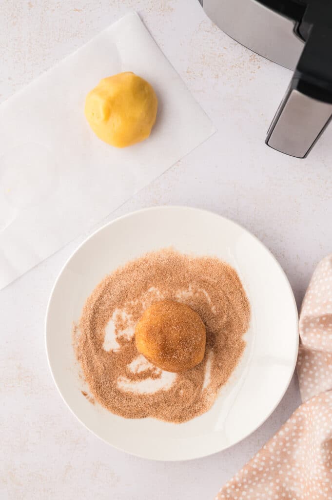 A portion of snickerdoodle cookie dough on a white plate is being coated with cinnamon and sugar.
