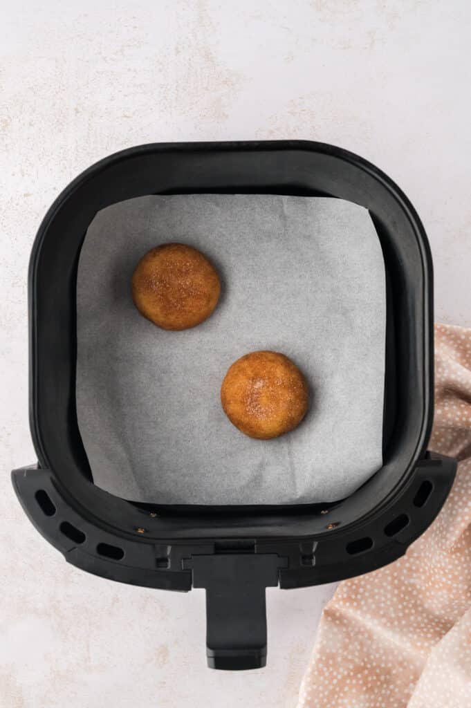 An air fryer basket lined with parchment paper holds two round, slightly flattened snickerdoodle cookie dough portions in the center.