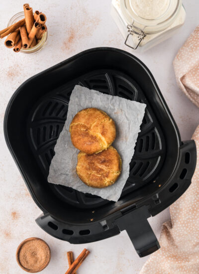 An overhead view of an air fryer basket holding two cooked snickerdoodle cookies, placed on a white countertop dusted with cinnamon and sugar around the basket.