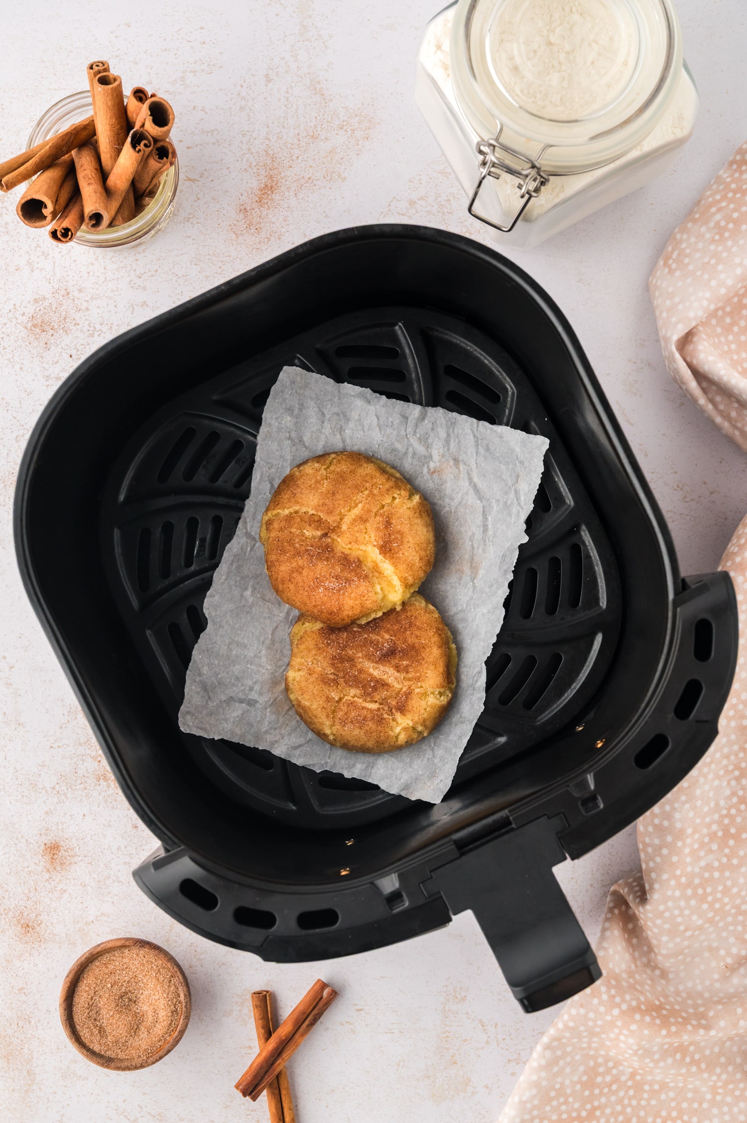 An overhead view of an air fryer basket holding two cooked snickerdoodle cookies, placed on a white countertop dusted with cinnamon and sugar around the basket.