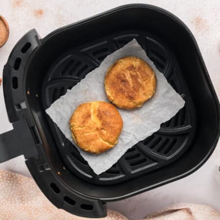 Two cooked snickerdoodle cookies sit inside an air fryer basket, surrounded by a ring of cinnamon sticks, seen from above.