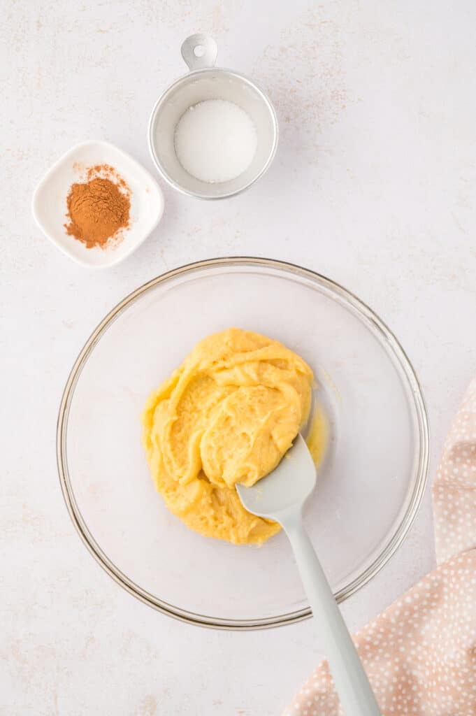 Snickerdoodle cookie dough is being mixed in a clear glass bowl, with a small dish of cinnamon and a small dish of sugar beside it.