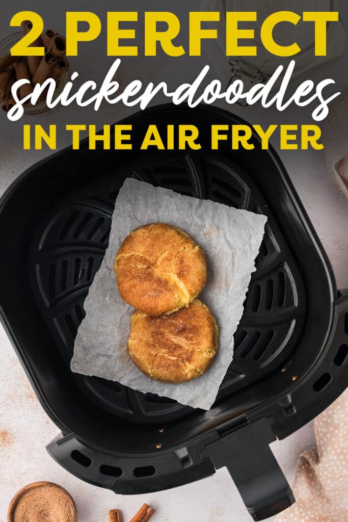An air fryer basket holds a small piece of parchment paper, with two cooked snickerdoodle cookies resting on top.