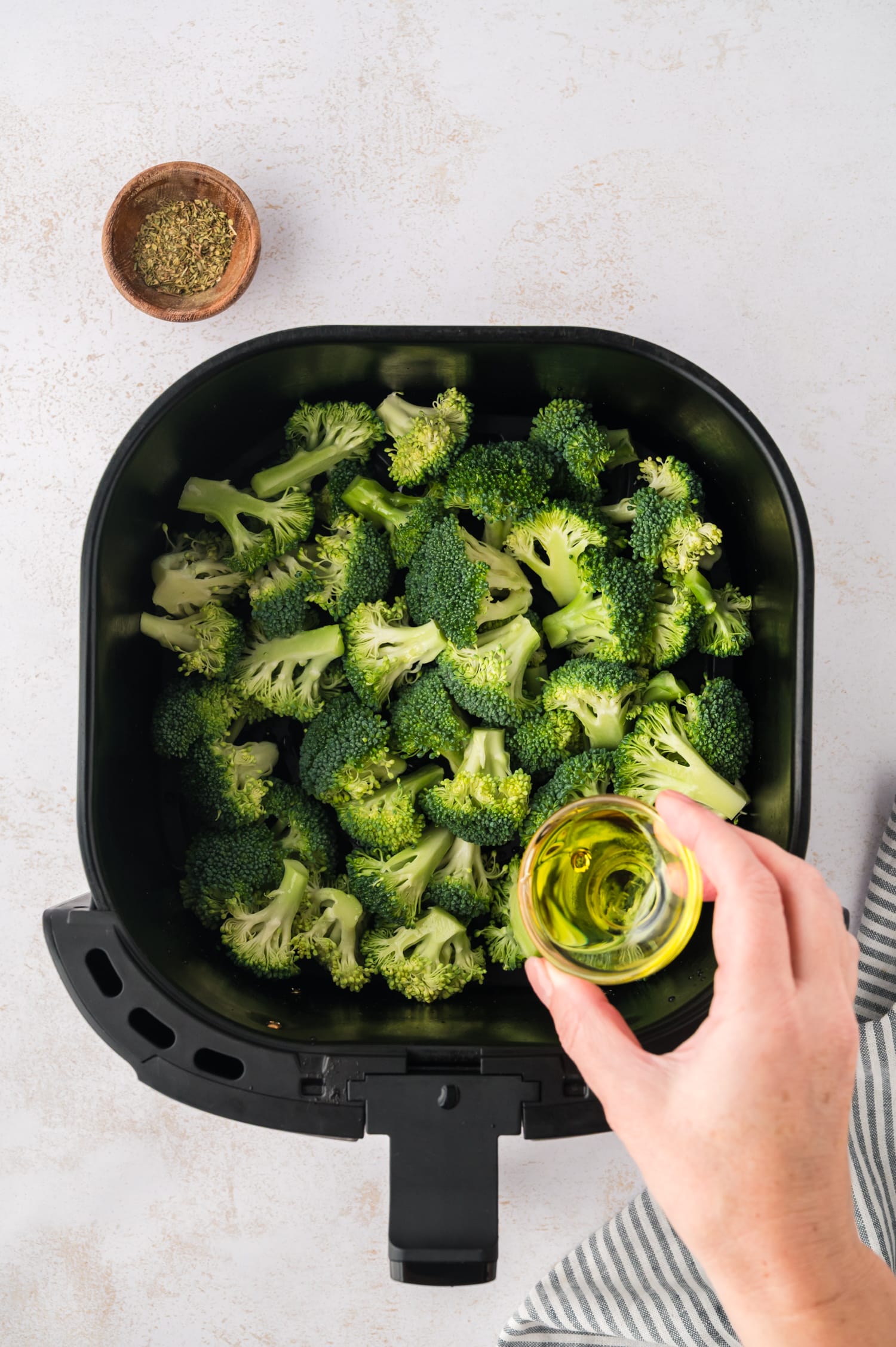 Raw broccoli and an air fryer basket with olive oil being held over it.