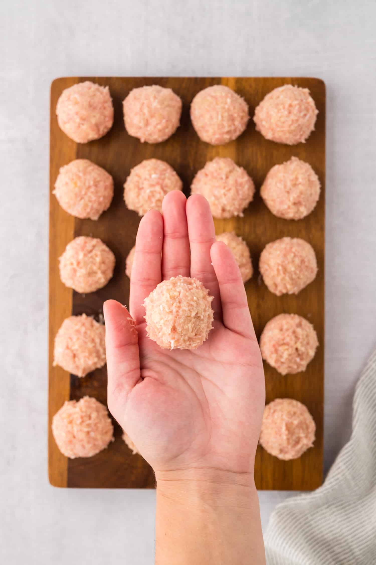Raw chicken meatballs on a wooden cutting board with a person holding one of the shaped meatballs.