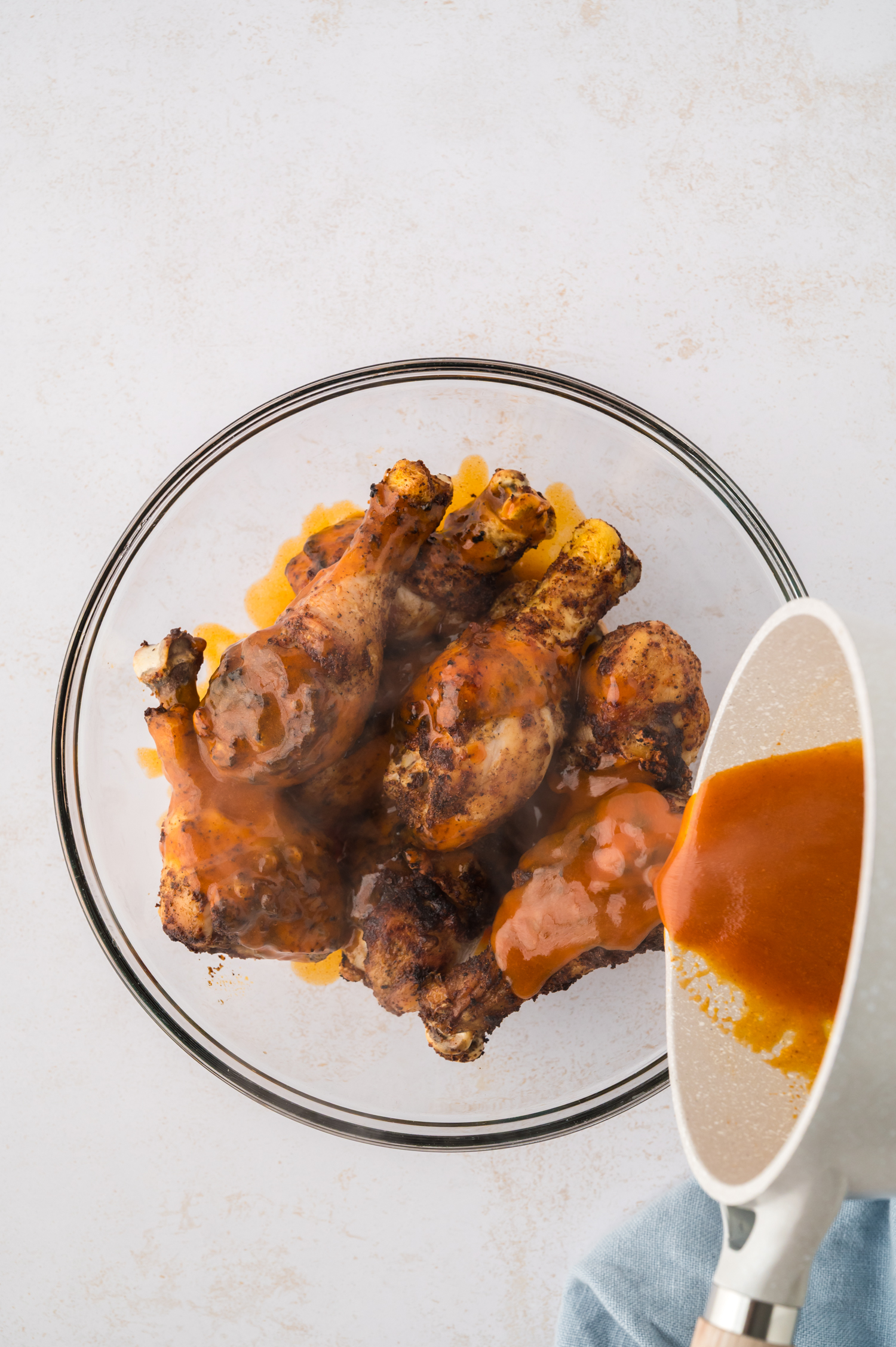 Buffalo sauce is being poured over cooked chicken drumsticks, coating their skin with a glossy, spicy layer.