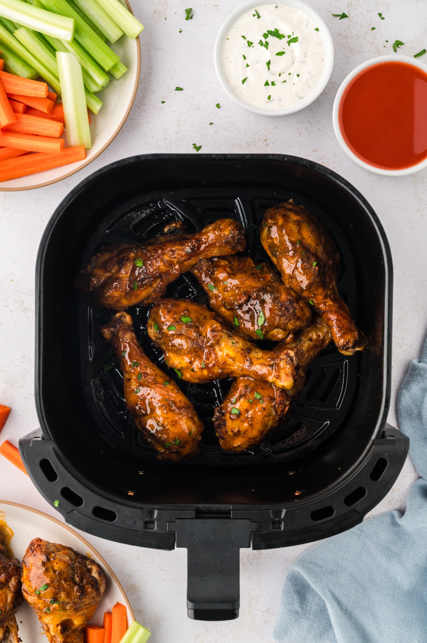 An overhead view of buffalo chicken drumsticks arranged in an air fryer basket, set beside a plate of sliced carrots and celery sticks.