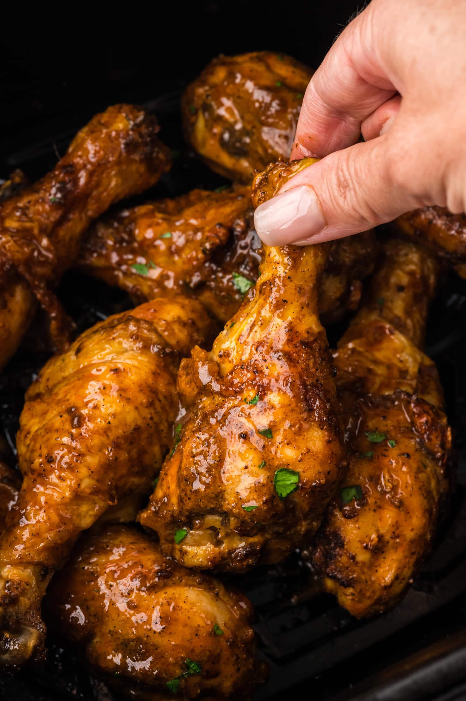 A woman reaches into an air fryer basket and lifts a buffalo chicken drumstick with her hand, grasping it by the bone as it comes up from the basket.