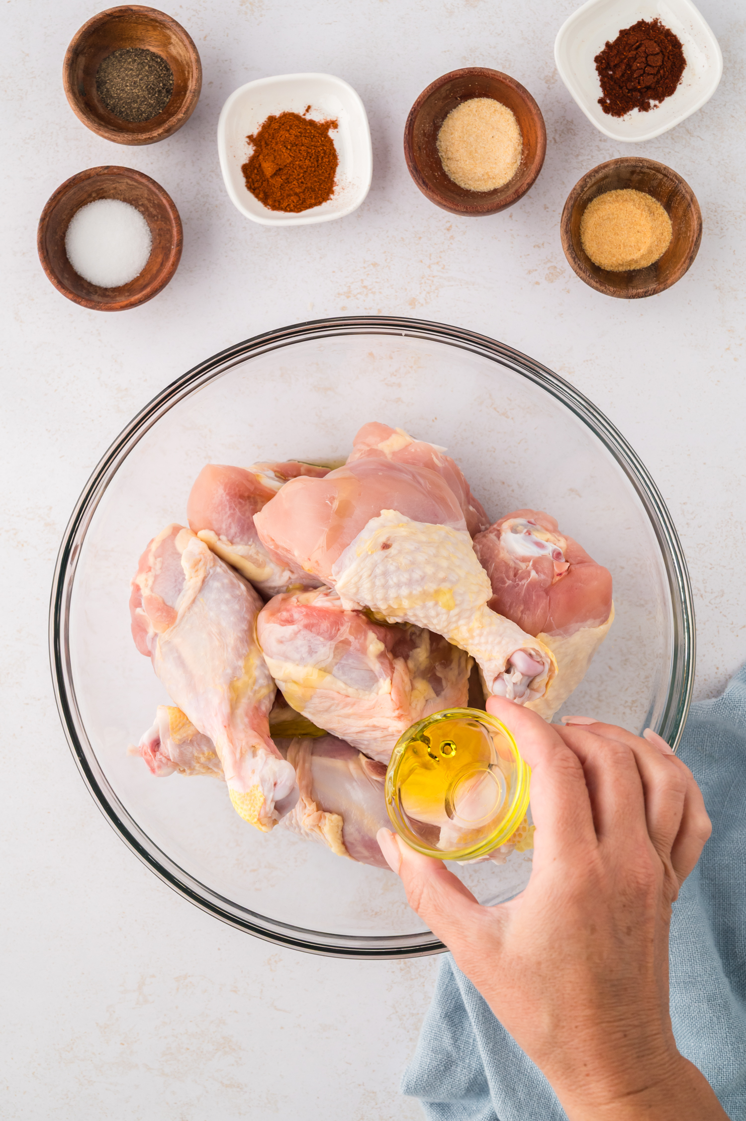 Raw chicken drumsticks sit in a mixing bowl as a woman pours olive oil over them, coating the skin before seasoning.