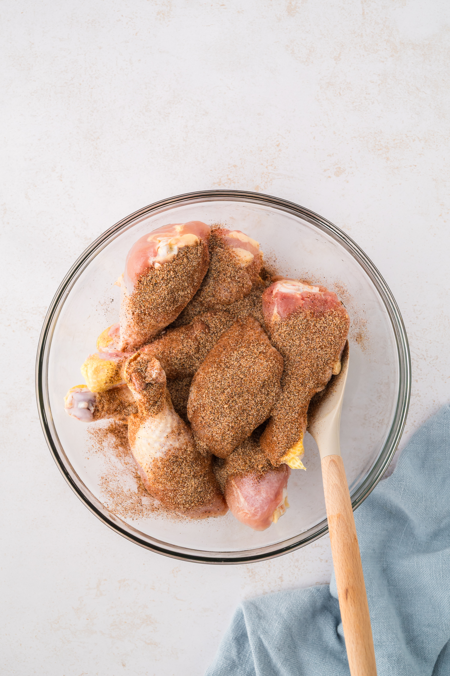 Raw chicken drumsticks are being tossed in seasoning inside a clear glass bowl, coating the skin evenly.