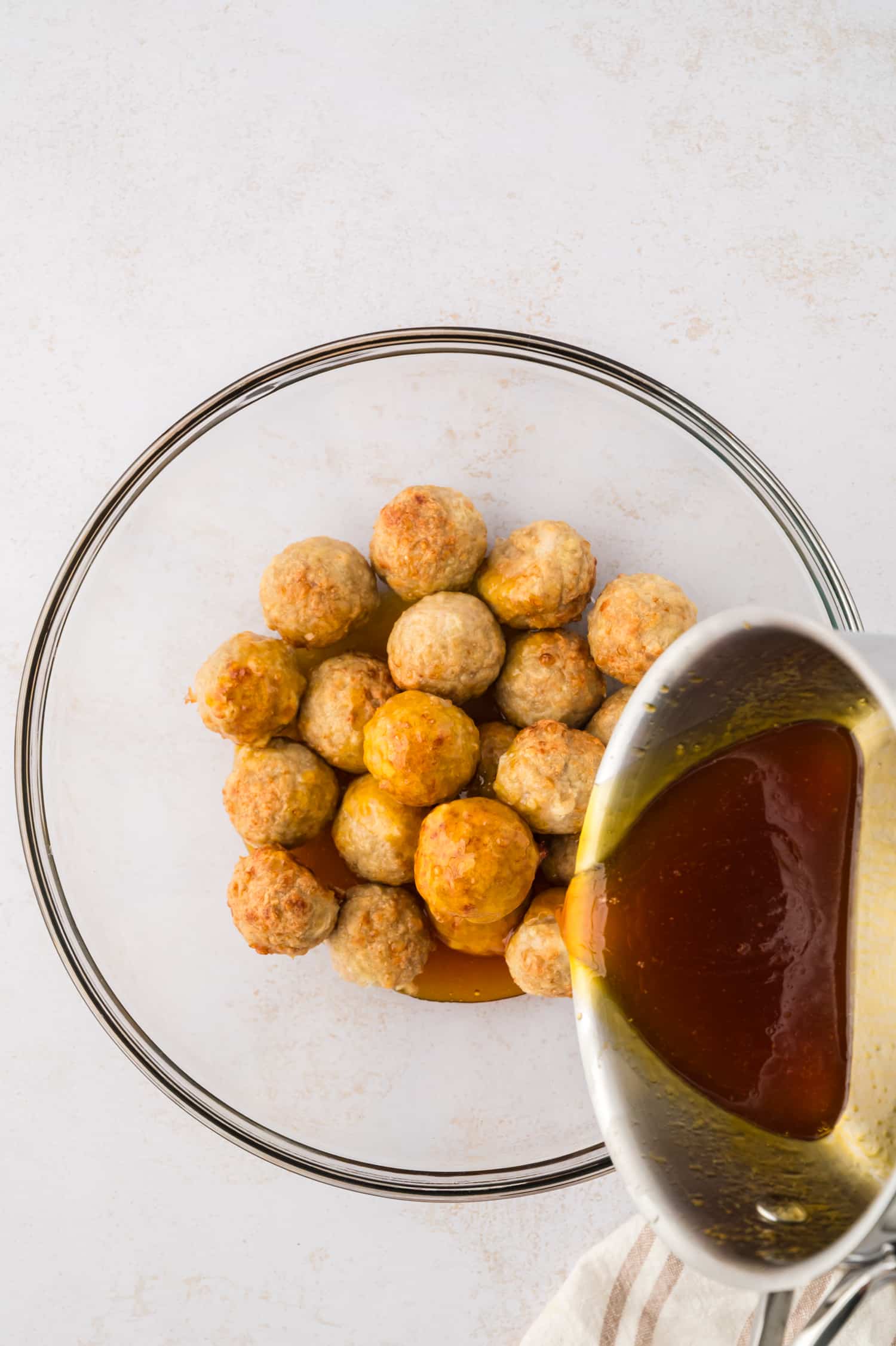Hot honey is being poured over cooked chicken meatballs in a mixing bowl, coating them with a glossy, sweet glaze.