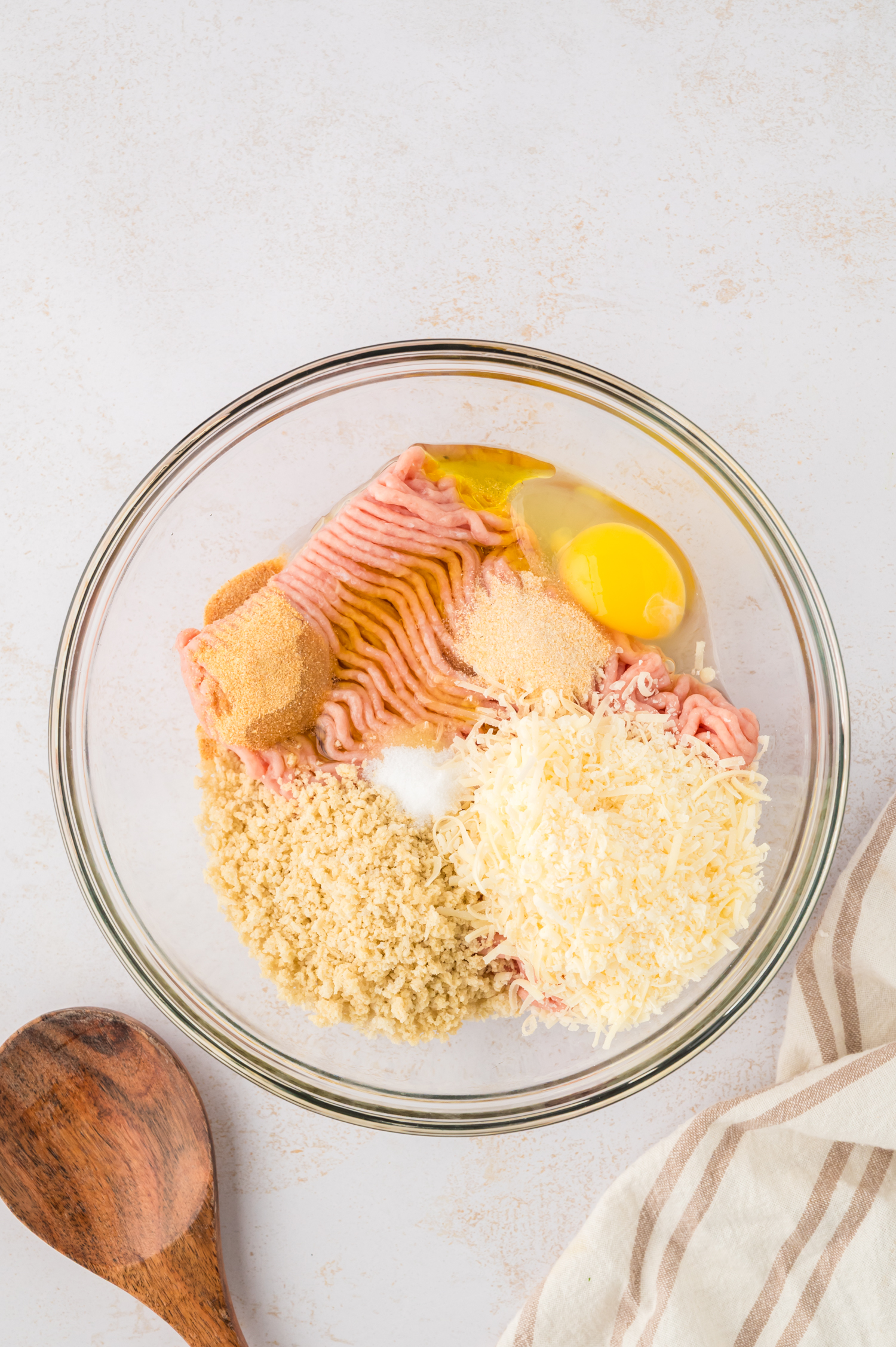 Raw ingredients for chicken meatballs are combined in a clear glass mixing bowl, ready to be mixed together.